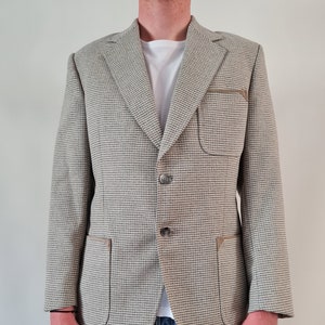 Könnte beinhalten: Ein beigefarbener Blazer mit einem braunen und weißen Karomuster. Der Blazer hat zwei Pattentaschen mit braunen Lederbesätzen und einen Knopfverschluss.