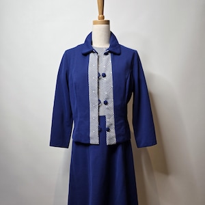 Może przedstawiać: Zestaw vintage z granatową spódnicą prezentowany na manekinie. Zestaw składa się z kurtki z kołnierzem z wzorzystym panelem i guzikami, połączonej z pasującą spódnicą do kolan. Kurtka ma długie rękawy. Wzorzysty panel jest biało-czarny.