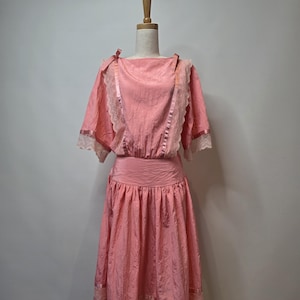 Puede incluir: Vestido rosa vintage con cuello barco y mangas cortas. El vestido presenta un ribete de encaje en las mangas y el dobladillo, una falda fruncida y un detalle de cinta en los hombros. Se exhibe en un maniquí.