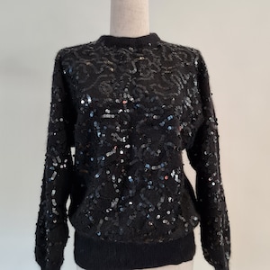 May include: Black sweater with long sleeves and a crew neck. The sweater is adorned with many black sequins in a swirling design across the front and sleeves. The cuffs and hem are ribbed.