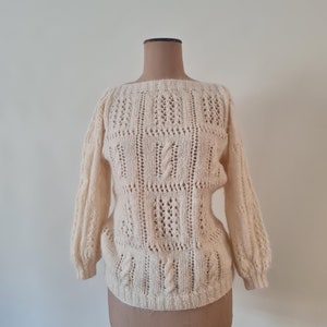May include: A white knitted jumper with a delicate openwork pattern. The jumper has a boat neck and long sleeves.