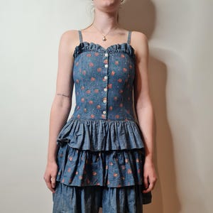 以下が含まれることがあります： 花柄のプリントが施された青いデニムのドレス。スカートはフリル付き。ドレスはフロントにボタン留め、細いストラップ付きです。