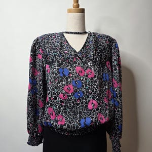 May include: A black blouse with a floral pattern in pink, blue, and white. The long-sleeved top has a V-neck and elastic cuffs and waist. The blouse is displayed on a mannequin.