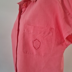 May include: Coral-colored short-sleeved shirt with a button-down front and a chest pocket. The pocket features an embroidered crest design. The shirt is made of a smooth fabric and has a classic collar.