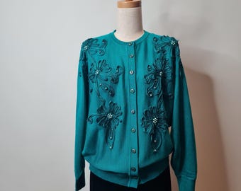 Pode incluir: Um cardigan de malha azul turquesa com fecho de botões e bordado floral. O bordado é feito com fio azul turquesa escuro e tem um design de contas.