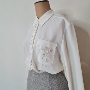 May include: A white long-sleeved blouse with a button-up collar and a floral embroidered pocket. The blouse is on a mannequin and paired with a grey skirt.