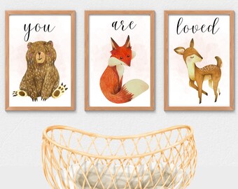 May include: Three framed art prints with woodland animal illustrations. The prints feature a bear, fox, and deer, with the words "you", "are", and "loved" in black script. A light brown wicker bassinet is in the foreground.