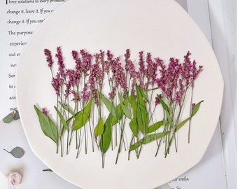 May include: A white, irregularly shaped plate decorated with dried purple flowers and green leaves. The plate is set on a white surface with text visible in the background. The flowers are arranged in a natural, organic style.