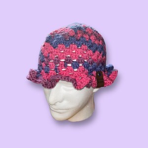 May include: A handmade crochet bucket hat in shades of pink, purple, and blue. The hat features a scalloped brim and a tag. The hat is displayed on a white mannequin head against a lavender background. The hat is a unique accessory.