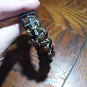 Pu&ograve; includere: Un braccialetto in paracord marrone e verde con perline di legno marrone e un ciondolo a forma di croce argentato.