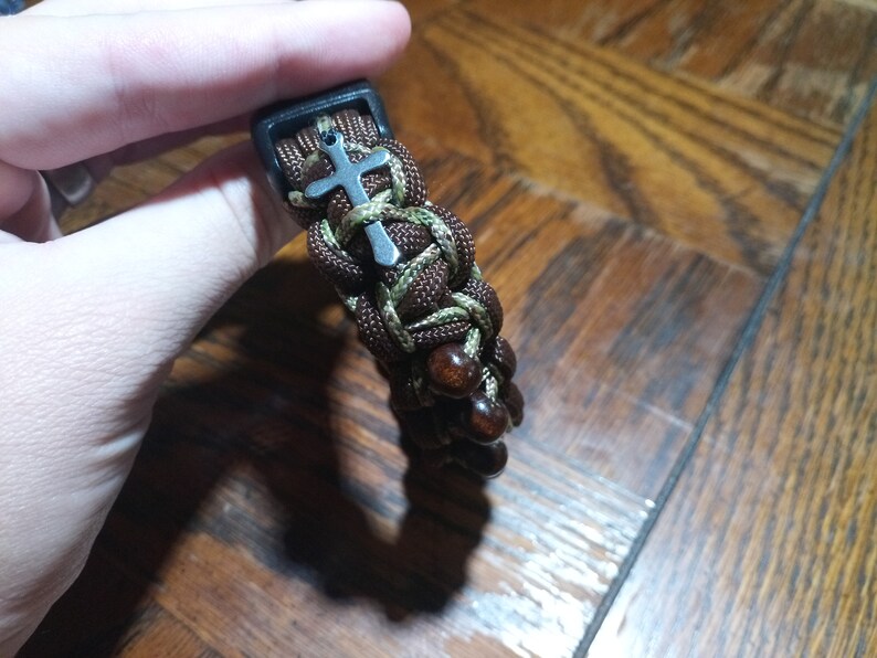 Pu&ograve; includere: Un braccialetto in paracord marrone e verde con perline di legno marrone e un ciondolo a forma di croce argentato.