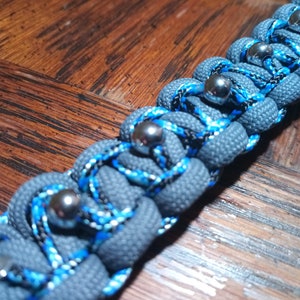 Pu&ograve; includere: Un braccialetto in paracord grigio con accenti blu e neri e perline metalliche argentate.