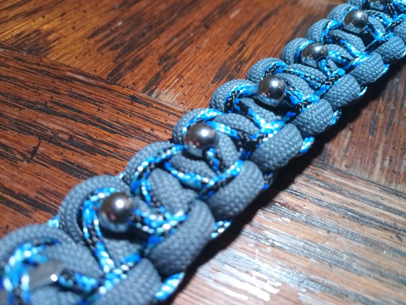 Pu&ograve; includere: Un braccialetto in paracord grigio con accenti blu e neri e perline metalliche argentate.