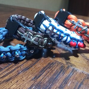 Pu&ograve; includere: Quattro braccialetti intrecciati con ciondoli a croce argentati e perline. I braccialetti sono blu, marroni, bianchi e arancioni.