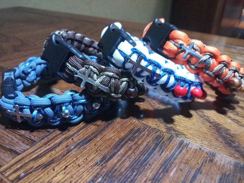 Pu&ograve; includere: Quattro braccialetti intrecciati con ciondoli a croce argentati e perline. I braccialetti sono blu, marroni, bianchi e arancioni.