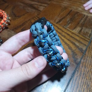 Pu&ograve; includere: Un braccialetto in paracord grigio e blu con un ciondolo a croce argentato e perline argentate.