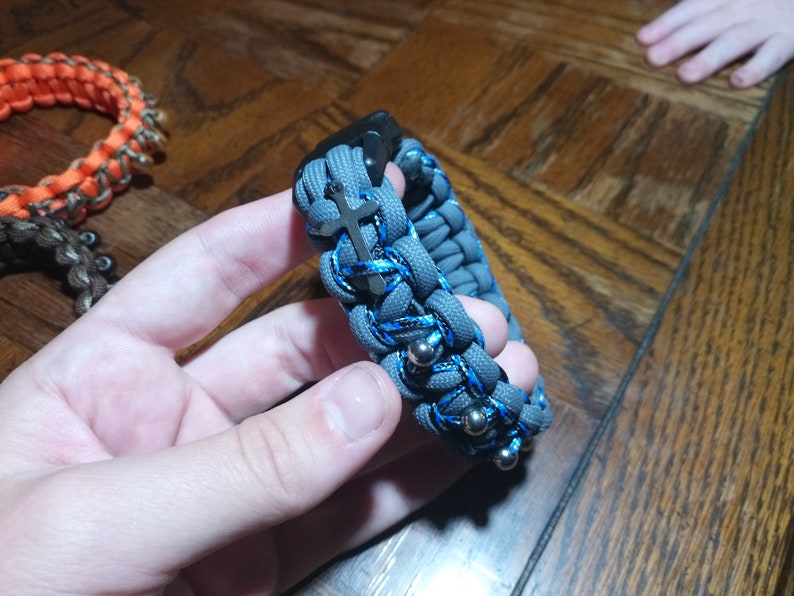 Pu&ograve; includere: Un braccialetto in paracord grigio e blu con un ciondolo a croce argentato e perline argentate.
