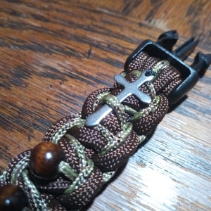 Pu&ograve; includere: Un braccialetto in paracord marrone e verde con un ciondolo a forma di croce argentato e una fibbia in plastica nera.