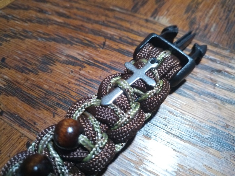 Pu&ograve; includere: Un braccialetto in paracord marrone e verde con un ciondolo a forma di croce argentato e una fibbia in plastica nera.