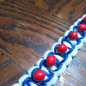 Pu&ograve; includere: Un braccialetto in paracord bianco e blu con perline rosse. Il braccialetto &egrave; intrecciato con un motivo complesso e ha una chiusura in metallo.