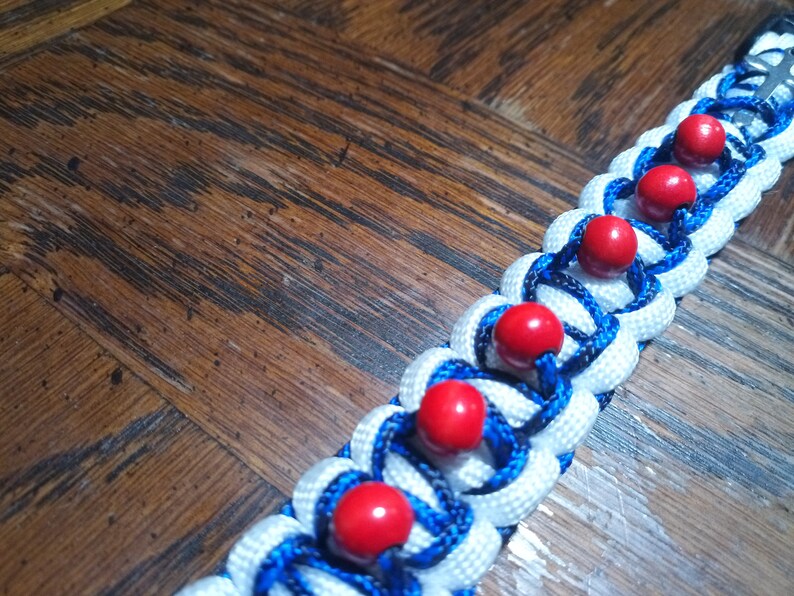 Pu&ograve; includere: Un braccialetto in paracord bianco e blu con perline rosse. Il braccialetto &egrave; intrecciato con un motivo complesso e ha una chiusura in metallo.