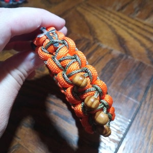 Pu&ograve; includere: Un braccialetto intrecciato con paracord arancione e verde, con perline di legno e una chiusura a croce argentata.
