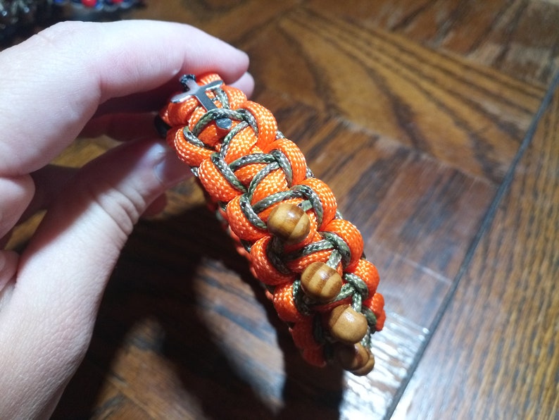 Pu&ograve; includere: Un braccialetto intrecciato con paracord arancione e verde, con perline di legno e una chiusura a croce argentata.