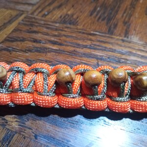 Pu&ograve; includere: Un braccialetto in paracord con cordoncino a motivo mimetico arancione e verde e perline di legno.