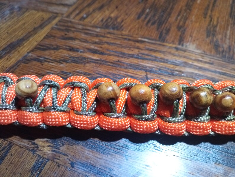 Pu&ograve; includere: Un braccialetto in paracord con cordoncino a motivo mimetico arancione e verde e perline di legno.