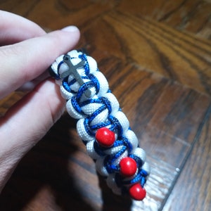 Pu&ograve; includere: Braccialetto in paracord bianco e blu con perline rosse e un ciondolo a forma di croce argentato.