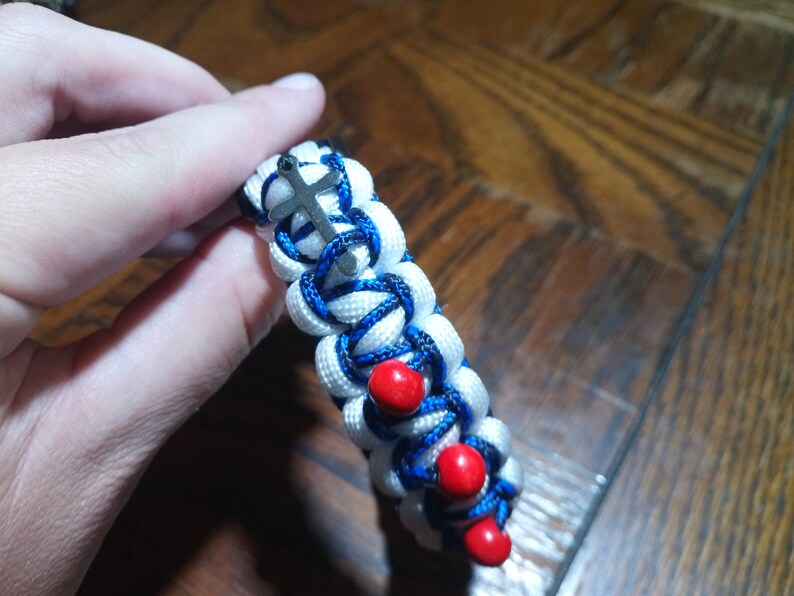 Pu&ograve; includere: Braccialetto in paracord bianco e blu con perline rosse e un ciondolo a forma di croce argentato.