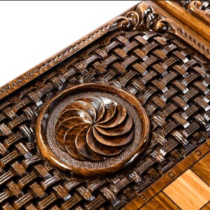 May include: A wooden chessboard with a decorative inlay of a woven pattern and a carved circular design. The chessboard is made of dark wood with light wood squares.