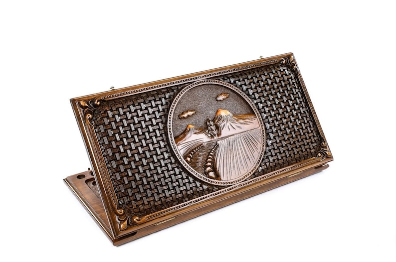 May include: A wooden game box with a carved design of a mountain landscape. The box has a woven pattern on the lid and is closed.