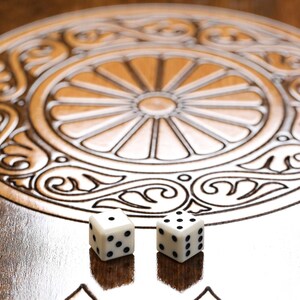 May include: Two white dice with black dots on a wooden game board with an intricate carved design. The dice are in the centre of the board.