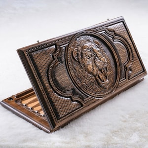 May include: A wooden backgammon set with a carved lion head design on the lid. The lid is open, revealing the game board and playing pieces.