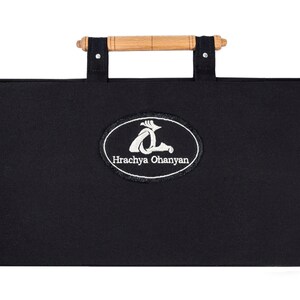 Puede incluir: Estuche de transporte de lona negra con asa de madera y un logotipo ovalado bordado en blanco que dice "Hrachya Ohanyan".