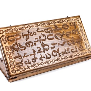以下が含まれることがあります： ヒンジ付きの蓋が付いた木製のゲームボックス。蓋は、アルメニア語のアルファベットの複雑なデザインで飾られています。ボックスは閉じており、白い背景の上に置かれています。
