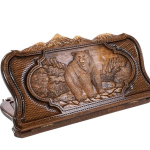 May include: A wooden game box with a carved bear design. The box is closed and has a hinged lid. The bear is brown and has a mountain scene behind it.