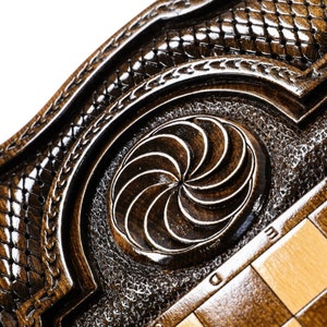 Puede incluir: Primer plano de un tablero de ajedrez de madera con un dise&ntilde;o geom&eacute;trico tallado en el centro. El tablero est&aacute; hecho de madera oscura con una incrustaci&oacute;n de madera clara. El tablero de ajedrez est&aacute; en un marco decorativo con un dise&ntilde;o tallado.