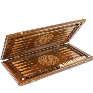 May include: A wooden backgammon set with an intricate design on the lid and board. The set includes 15 brown and 15 white playing pieces.