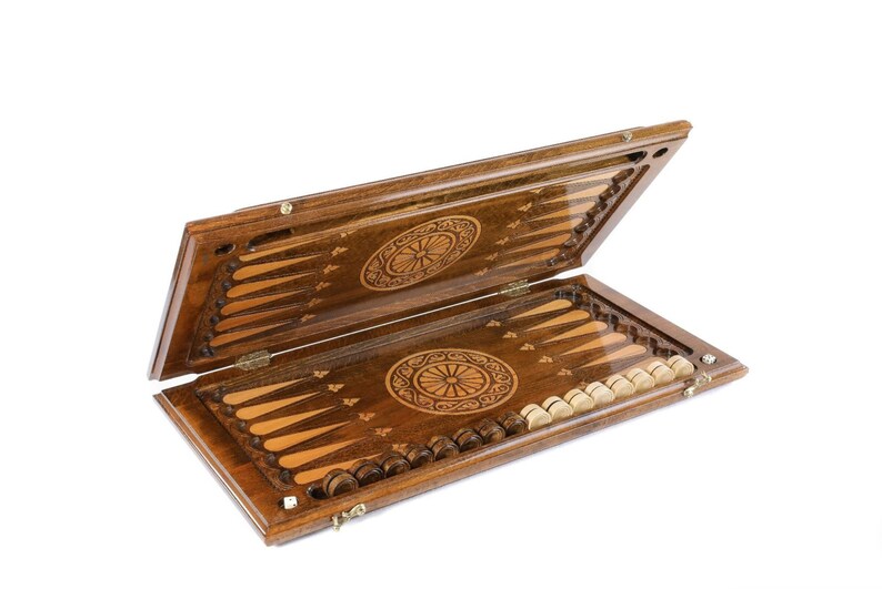 May include: A wooden backgammon set with an intricate design on the lid and board. The set includes 15 brown and 15 white playing pieces.