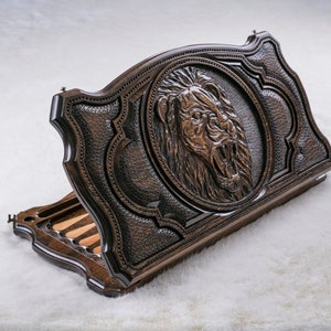 May include: A wooden backgammon game set with a lion carved into the lid. The lid is open and the game pieces are visible inside.
