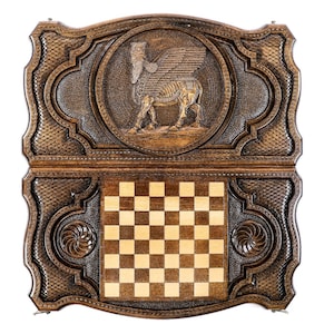 Puede incluir: Un tablero de ajedrez de madera con intrincados tallados. La parte superior del tablero presenta una imagen tallada de un toro alado. El tablero de ajedrez est&aacute; cerrado y la superficie de juego es visible.