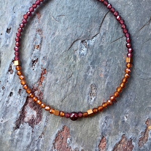 Könnte beinhalten: Ein Armband aus Granat- und Bernsteinperlen mit goldenen Akzenten. Das Armband besteht aus kleinen, runden Perlen in Rot- und Orangetönen, die mit goldenen quadratischen Perlen durchsetzt sind.