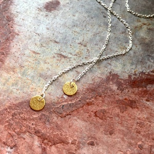 Könnte beinhalten: Ohrringe mit silbernen Ketten und kleinen, strukturierten, goldfarbenen, kreisförmigen Anhängern. Die Ohrringe werden vor einem marmorierten, rötlich-braunen Steinhintergrund präsentiert, was einen Kontrast erzeugt.