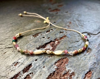 May include: A delicate bracelet made with a variety of colorful beads, including pink, green, and white. The bracelet is adjustable with a sliding knot and features gold accents.