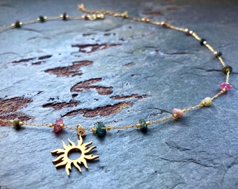 May include: A delicate gold necklace featuring a sun pendant and colorful gemstone beads. The necklace has a fine chain with small, multi-colored beads and a gold sun charm. The gemstones include pink, green, and blue stones.