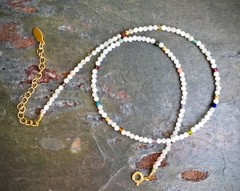 Könnte beinhalten: Eine Halskette aus weißen Edelsteinen mit einer goldenen Kette und einem Verschluss. Die Halskette besteht aus kleinen, runden Perlen, in die einige farbige Perlen eingestreut sind.