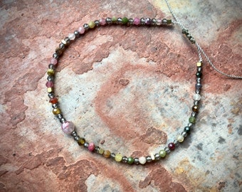 May include: A delicate necklace featuring a strand of small, faceted beads in various colors, including pink, green, yellow, and gray. The beads are interspersed with metallic accents and strung on a thin, silver-colored cord. The necklace is displayed against a textured, reddish-brown surface.
