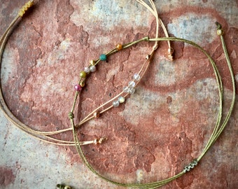 May include: Two delicate bracelets with colorful beads and gold accents. The bracelets are made of thin, natural-colored cord and green cord. The beads are a mix of clear, green, pink, and gold.